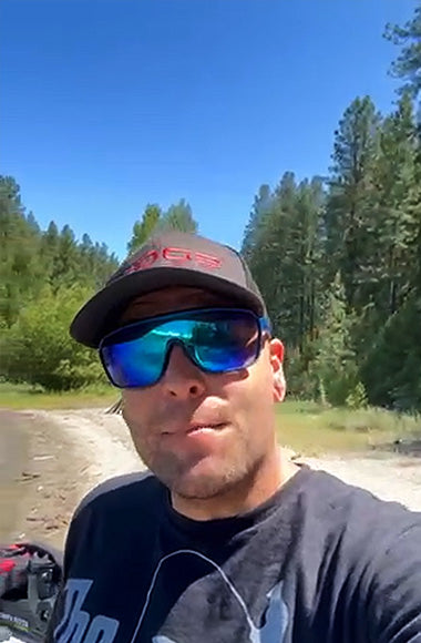 Man wearing sunglasses and cap in a forested area near a lake.